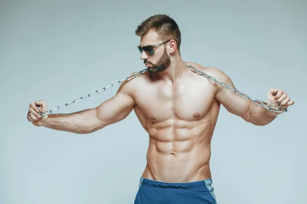 .fisiculturista posando. Bonito desportista poder masculino. Fitness musculado em calções azuis e óculos de sol. sobre fundo cinzento isolado. Homem com tronco musculado. Torso forte do modelo da aptidão do homem atlético — Fotografia de Stock