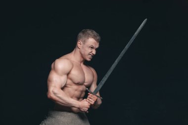 Vücut geliştirmeci adam siyah arka plan üzerine izole bir kılıç ile poz. Ciddi gömleksiz adam mascular vücudunu gösteren