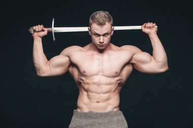 Vücut geliştirmeci adam siyah arka plan üzerine izole bir kılıç ile poz. Ciddi gömleksiz adam mascular vücudunu gösteren