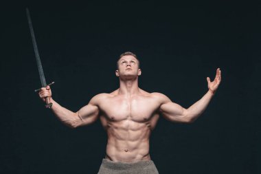 Vücut geliştirmeci adam siyah arka plan üzerine izole bir kılıç ile poz. Ciddi gömleksiz adam mascular vücudunu gösteren