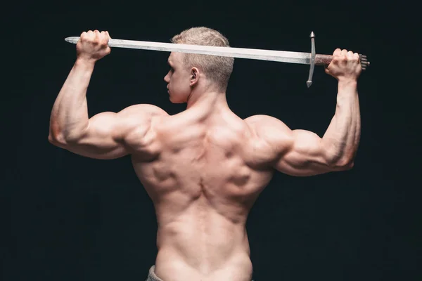 Bodybuilder homme posant avec une épée isolée sur fond noir. Sérieux homme torse nu démontrant son corps masqué. épée pour tête — Photo