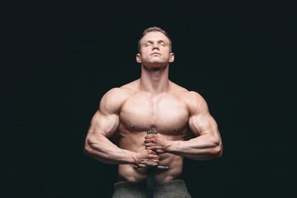 Bodybuilder Mann posiert mit einem Schwert isoliert auf schwarzem Hintergrund. ernster, hemdsloser Mann, der seinen maskulinen Körper demonstriert. Hände auf einem Schwert — Stockfoto