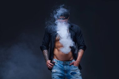 Vaper. Adam mavi kot pantolon giymiş, siyah gömlek ve siyah beyzbol şapkası dövmeler ile koyu arka plan üzerinde elektronik sigara duman