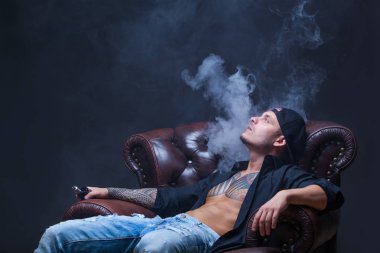Vaper. Dövmeli adam koyu arka plan üzerinde elektronik sigara duman deri kanepede oturuyor.