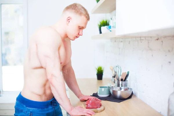 Hombre musculoso con un torso desnudo en la cocina con un trozo de carne de res, el concepto de una dieta saludable. Estilo de vida atlético . — Foto de Stock