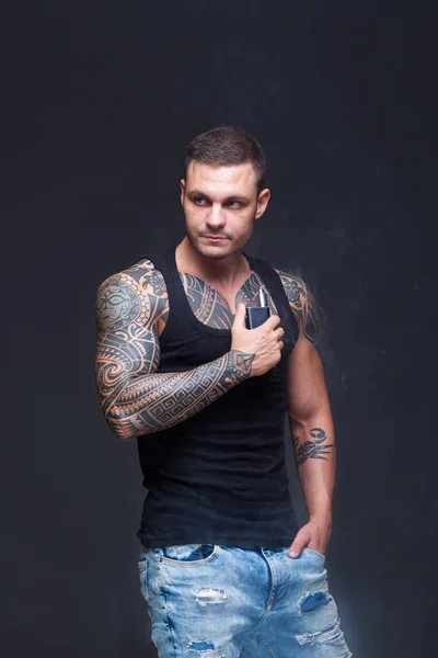 Vaper. The man with a muscular torso with tattoos smoke an electronic cigarette on the dark background — Stock Photo, Image