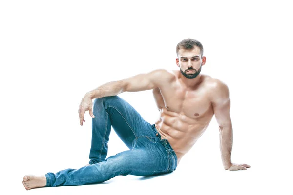 Fisiculturista posando. Bonito desportista poder masculino. Fitness musculado em jeans azuis. sobre fundo branco isolado. senta-se no chão — Fotografia de Stock