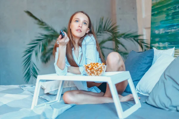 Hermosa joven sonriente mujer viendo una película en la cama y comiendo palomitas de maíz . — Foto de Stock