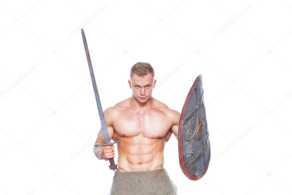 Bodybuilder man posing with a sword and shield isolated on white background. Serious shirtless man demonstrating his mascular body.