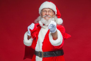 Noel. Noel Baba hediye onun arkasında bir torba ile beyaz eldiven gülümseyen onun parmağı kameraya çekiyor. Kırmızı zemin üzerine izole.