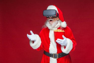 Noel. Noel Baba kırmızı bir kova patlamış mısır tutarak siyah sanal gerçeklik bardaklarda. Sanal gerçeklik bir film izlerken. Kırmızı zemin üzerine izole.