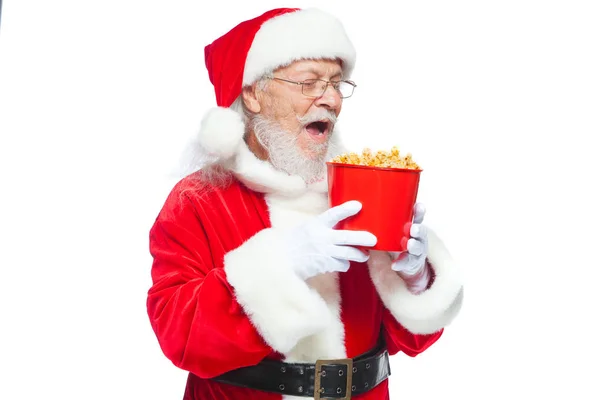 Natale. Babbo Natale in guanti bianchi con la bocca aperta tiene un secchio rosso di popcorn e si prepara a mangiarlo. Il concetto di visitare un cinema, guardare un film con i popcorn. Isolato su bianco — Foto Stock