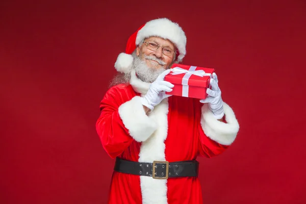 Weihnachten. Der freundliche Weihnachtsmann in weißen Handschuhen hält eine rote Geschenkschachtel mit einer Schleife am Ohr. hört er zu. isoliert auf rotem Hintergrund. — Stockfoto