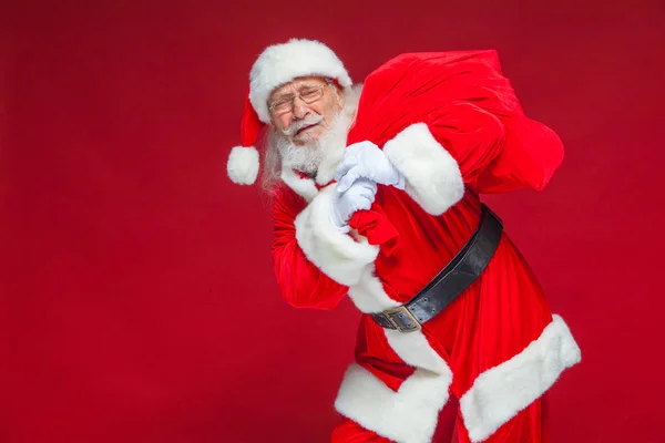 Weihnachten. Der freundliche und müde Weihnachtsmann in weißen Handschuhen trägt eine rote Tasche mit Geschenken über der Schulter. isoliert auf rotem Hintergrund. — Stockfoto