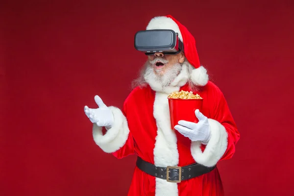 Christmas. Santa Claus in black virtual reality glasses holding a red bucket of popcorn. Watching a movie in virtual reality. Isolated on red background.