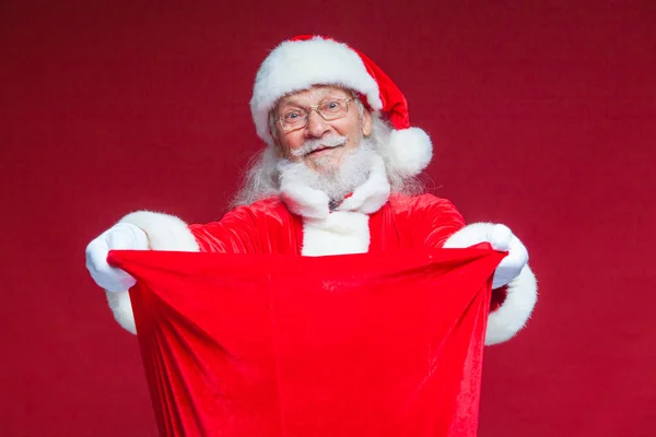 Christmas. Santa Claus in white gloves opened the bag with gifts and looks into it. Offers to take the gifts. Isolated on red background. — Stock Photo, Image