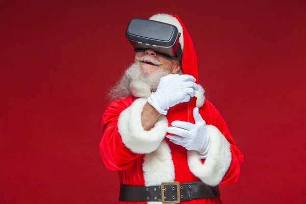 Noël. Le Père Noël en lunettes de réalité virtuelle noires tenant un seau rouge de pop-corn. Regarder un film en réalité virtuelle. Isolé sur fond rouge . — Photo
