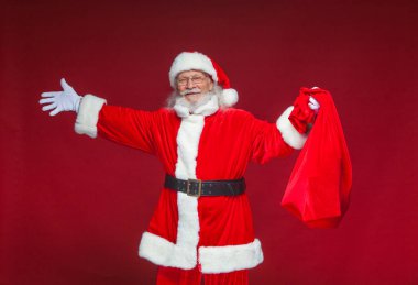 Noel. Nasıl Noel Baba gülümseyerek ellerini yanlara yayıldı. Kırmızı çanta hediye ile bir elinde tutuyor. Kırmızı zemin üzerine izole.