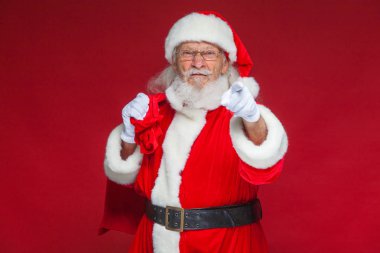 Noel. Noel Baba hediye arkasından bir torba ile memnun yüz ifadesi ile beyaz eldiven onun parmağı kameraya çekiyor. Kırmızı zemin üzerine izole.