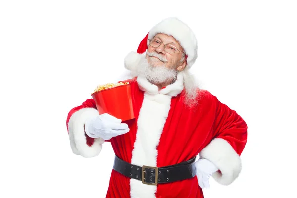 Navidad. Santa Claus sonriente y amable con guantes blancos sostiene un cubo rojo con palomitas de maíz con una mano, y pone la otra mano en su cinturón. El concepto de visitar el cine, ver una película con — Foto de Stock