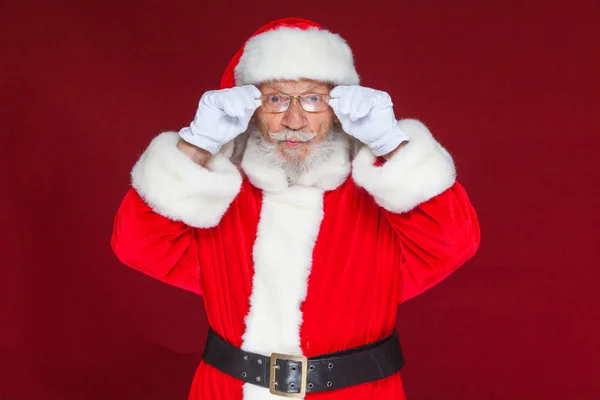 Weihnachten. Ernster Weihnachtsmann in weißen Handschuhen richtet seine Brille ein und starrt in die Kamera. isoliert auf rotem Hintergrund. — Stockfoto