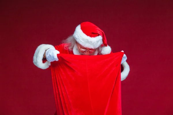 Weihnachten. Santa Claus in weißen Handschuhen öffnete die Tasche mit Geschenken und schaut hinein. bietet an, die Geschenke anzunehmen. isoliert auf rotem Hintergrund. — Stockfoto