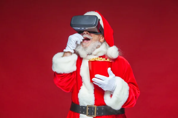 Noël. Le Père Noël en lunettes de réalité virtuelle noires tenant un seau rouge de pop-corn. Regarder un film en réalité virtuelle. Isolé sur fond rouge . — Photo