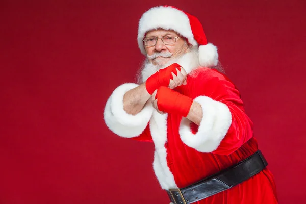 Weihnachten. Weihnachtsmann mit roten Bandagen, die sich um seine Hände wickeln, um Tritte zu imitieren. Kickboxen, Karate, Boxen. isoliert auf rotem Hintergrund. — Stockfoto