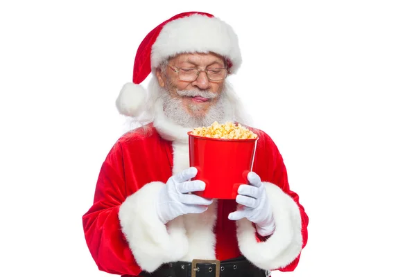 Natale. Babbo Natale in guanti bianchi tiene un secchio rosso di popcorn, lecca le labbra e si prepara a mangiarlo. Il concetto di visitare il cinema, guardare un film con i popcorn. Isolato su bianco — Foto Stock