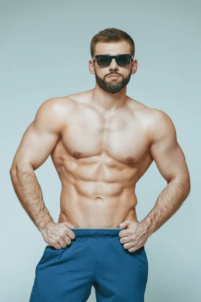 .fisiculturista posando. Bonito desportista poder masculino. Fitness musculado em calções azuis e óculos de sol. sobre fundo cinzento isolado. Homem com tronco musculado. Torso forte do modelo da aptidão do homem atlético — Fotografia de Stock