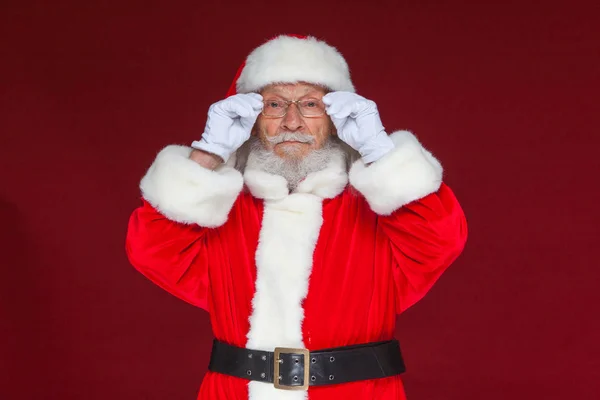 Weihnachten. Ernster Weihnachtsmann in weißen Handschuhen richtet seine Brille ein und starrt in die Kamera. isoliert auf rotem Hintergrund. — Stockfoto