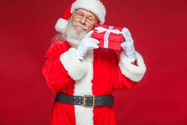 Weihnachten. Der freundliche Weihnachtsmann in weißen Handschuhen hält eine rote Geschenkschachtel mit einer Schleife am Ohr. hört er zu. isoliert auf rotem Hintergrund. — Stockfoto