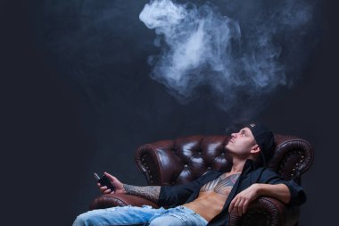 Vaper. Dövmeli adam koyu arka plan üzerinde elektronik sigara duman deri kanepede oturuyor.