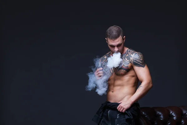 Vaper. El hombre con un torso desnudo muscular con tatuajes fuma un cigarrillo electrónico en el fondo oscuro — Foto de Stock