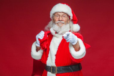 Noel. Noel Baba hediye onun arkasında bir torba ile beyaz eldiven gülümseyen onun parmağı kameraya çekiyor. Kırmızı zemin üzerine izole.
