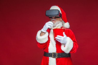 Noel. Noel Baba kırmızı bir kova patlamış mısır tutarak siyah sanal gerçeklik bardaklarda. Sanal gerçeklik bir film izlerken. Kırmızı zemin üzerine izole.