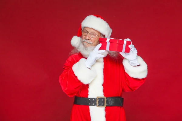 Weihnachten. Der freundliche Weihnachtsmann in weißen Handschuhen hält eine rote Geschenkschachtel mit einer Schleife am Ohr. hört er zu. isoliert auf rotem Hintergrund. — Stockfoto
