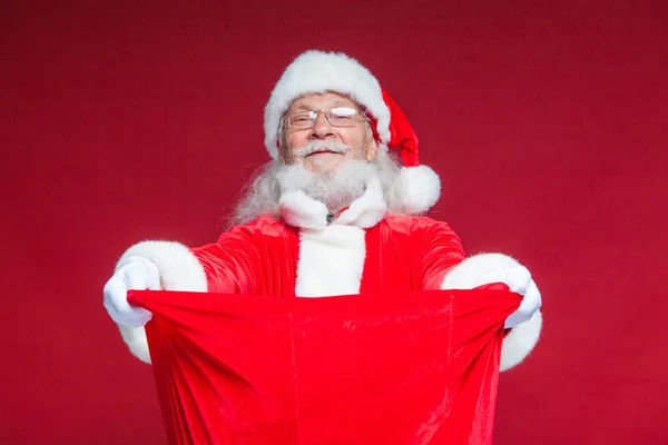 Weihnachten. Santa Claus in weißen Handschuhen öffnete die Tasche mit Geschenken und schaut hinein. bietet an, die Geschenke anzunehmen. isoliert auf rotem Hintergrund. — Stockfoto