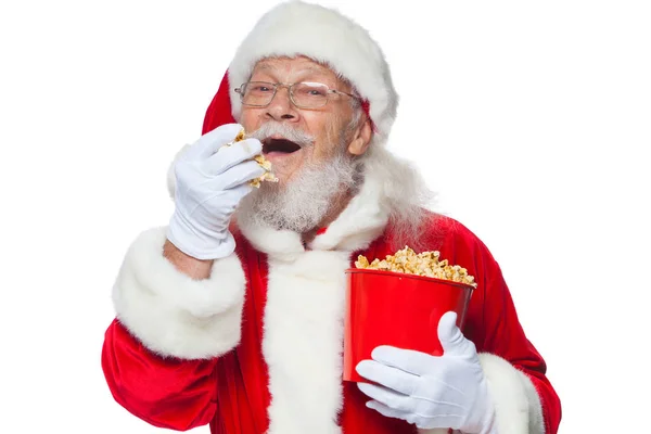 Natale. Sorridente, gentile Babbo Natale in guanti bianchi con la bocca aperta tiene un secchio rosso con popcorn con una mano, prende un po 'di popcorn e si prepara a mangiarlo con la seconda mano. Il concetto di — Foto Stock