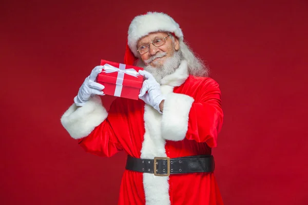Weihnachten. Der freundliche Weihnachtsmann in weißen Handschuhen hält eine rote Geschenkschachtel mit einer Schleife am Ohr. hört er zu. isoliert auf rotem Hintergrund. — Stockfoto