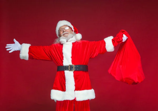 Weihnachten. Der freundlich lächelnde Weihnachtsmann breitete seine Hände zur Seite aus. In der einen Hand hält er eine rote Tüte mit Geschenken. isoliert auf rotem Hintergrund. — Stockfoto