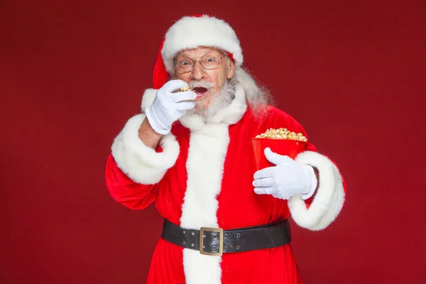 Christmas. Smiling, kind Santa Claus in white gloves with his mouth open holds a red bucket with popcorn with one hand, takes some popcorn and prepares to eat it with the second hand. The concept of — Stock Photo, Image