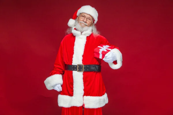 Weihnachten. Lächelnder Weihnachtsmann in weißen Handschuhen hält eine rote Geschenkschachtel mit Schleife in der Hand. zeigt auf das Geschenk. isoliert auf rotem Hintergrund. — Stockfoto