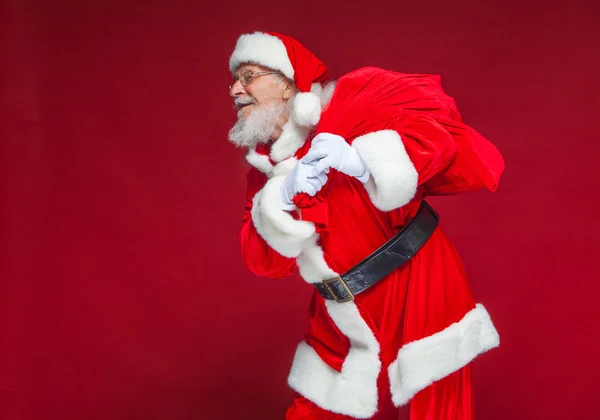 Weihnachten. Der freundliche und müde Weihnachtsmann in weißen Handschuhen trägt eine rote Tasche mit Geschenken über der Schulter. isoliert auf rotem Hintergrund. — Stockfoto