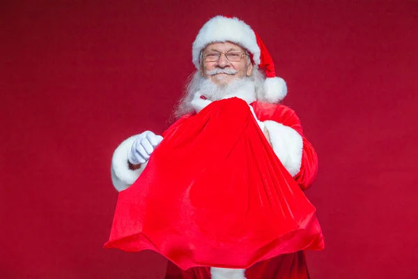 Christmas. A kind smiling Santa Claus is holding a red bag with presents in front of him embracing it. Isolated on red background. — Stock Photo, Image
