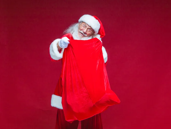 Christmas. Santa Claus in white gloves opened the bag with gifts and looks into it. Offers to take the gifts. Isolated on red background. — Stock Photo, Image