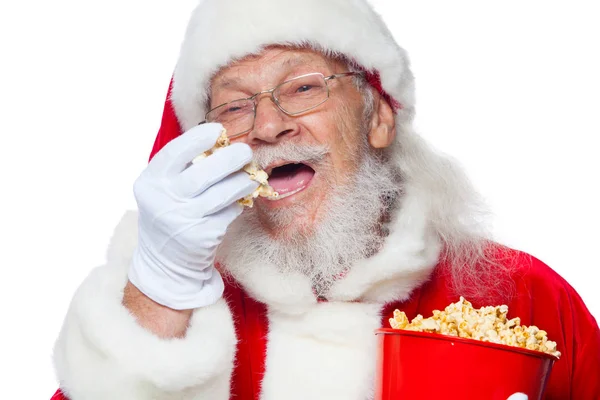 Natale. Sorridente, gentile Babbo Natale in guanti bianchi con la bocca aperta tiene un secchio rosso con popcorn con una mano, prende un po 'di popcorn e si prepara a mangiarlo con la seconda mano. Il concetto di — Foto Stock