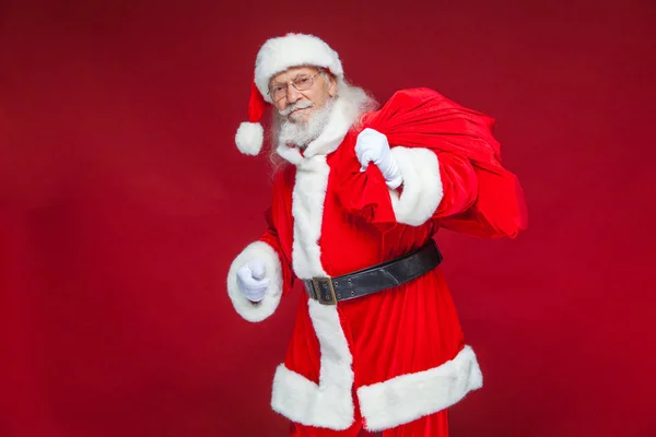 Weihnachten. Der freundliche und müde Weihnachtsmann in weißen Handschuhen trägt eine rote Tasche mit Geschenken über der Schulter. isoliert auf rotem Hintergrund. — Stockfoto