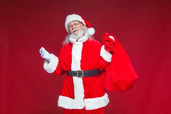 Weihnachten. Der freundlich lächelnde Weihnachtsmann breitete seine Hände zur Seite aus. In der einen Hand hält er eine rote Tüte mit Geschenken. isoliert auf rotem Hintergrund. — Stockfoto