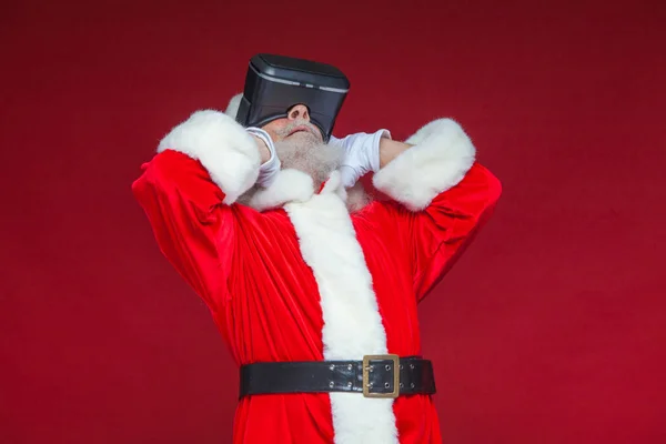 Weihnachten. Weihnachtsmann mit schwarzer Virtual-Reality-Brille macht Gesten mit den Händen. Überraschung, Emotion. Neue Technologien. isoliert auf rotem Hintergrund. — Stockfoto